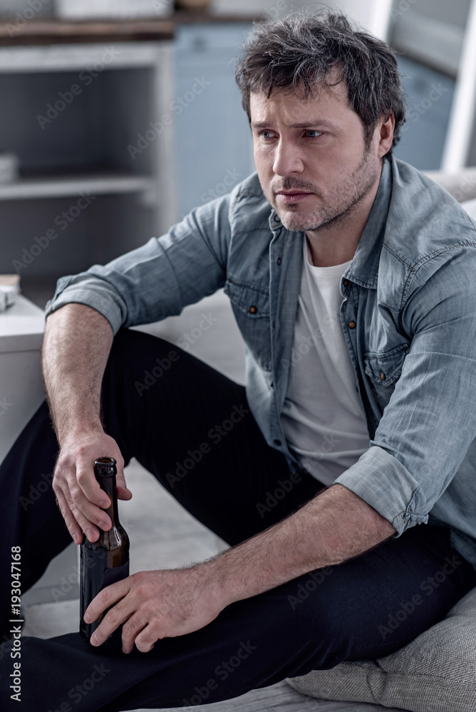Broken life. Uncombed unshaved man sitting on the floor with a bottle of beer in his hand and feeling awful while suffering from alcoholism