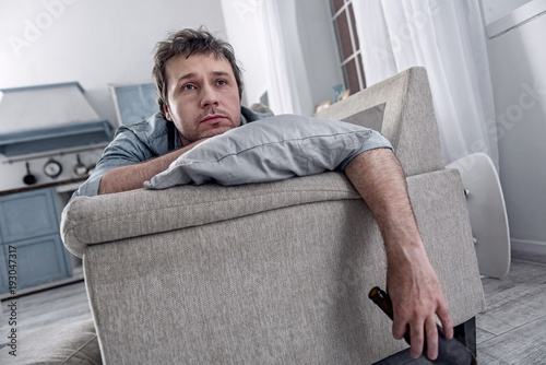 Bored person. Gloomy bored unemployed man spending his average day alone and feeling indifferent while resting on the sofa with a bottle of beer in his hand