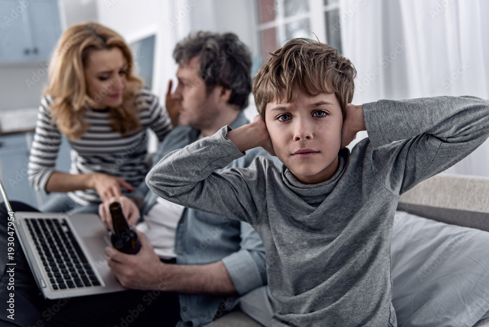 Tired child. Calm tired little boy feeling unhappy and closing his ears while his emotional rude parents having a quarrel near Tired child. Calm tired little boy feeling unhappy and closing his ears while his emotional rude parents having a quarrel near