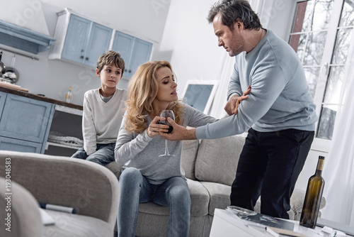 Rejecting addiction. Emotional serious worried man looking at his alcohol addicted wife and insisting on consulting a doctor while trying to take a glass of wine from her hand with their son watching