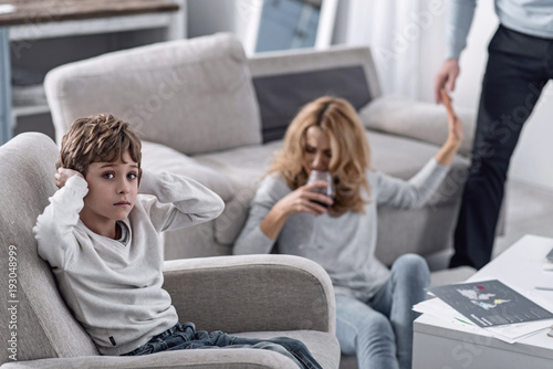 No endurance. Worried scared little boy closing his ears and feeling unhappy while his emotional alcoholic mother drinking wine on the floor and rejecting her addiction