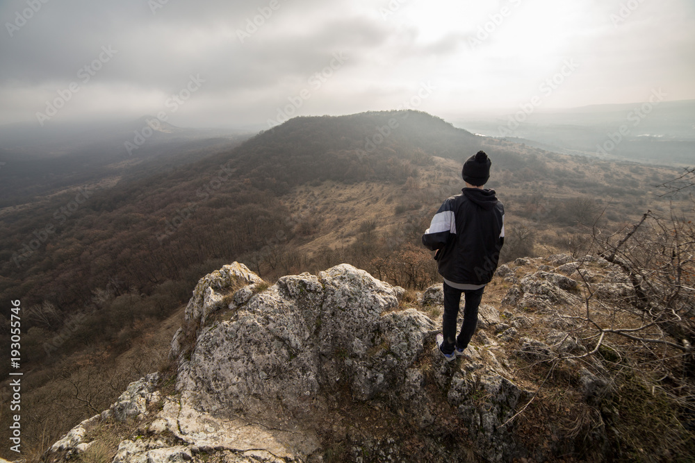 Wide angle panorama from a cliff on the top of a mountain with young man in back light sky.