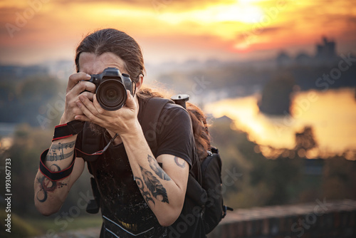 Young photographer with long hair and alternative style taking photographs with his dslr camera, capturing landscape and sunset in a park © Teodor Lazarev