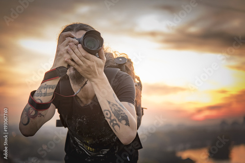 Young photographer with long hair and alternative style taking photographs with his dslr camera, capturing landscape and sunset in a park © Teodor Lazarev