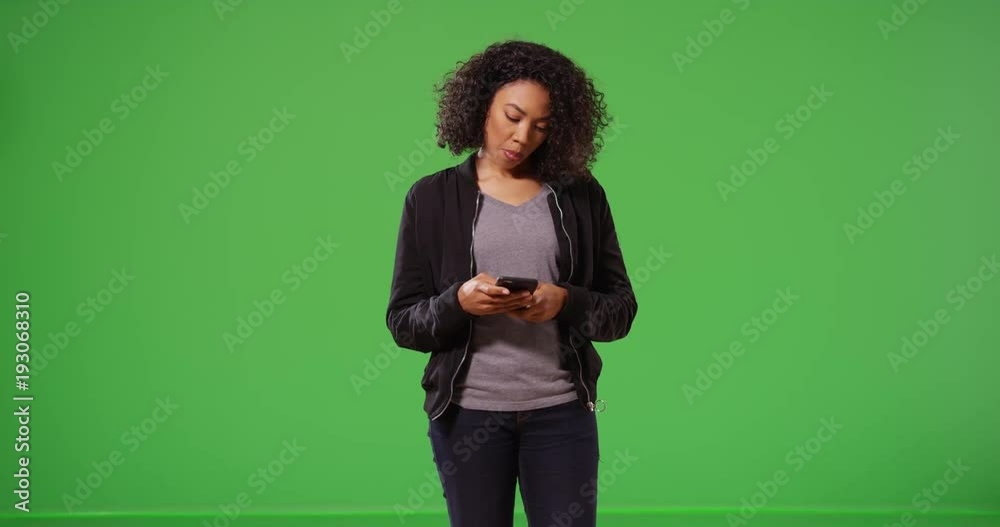 Woman walks on screen checks her mobile phone then exits off on greenscreen. Black millennial girl wearing bomber jacket receiving text messages on green background for keying or compositing. 4k