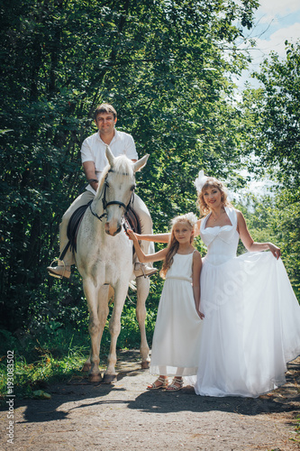 Man, woman dressed as a bride, girl and white horse in the park among the green trees © keleny