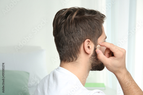 Young man putting hearing aid in ear indoors