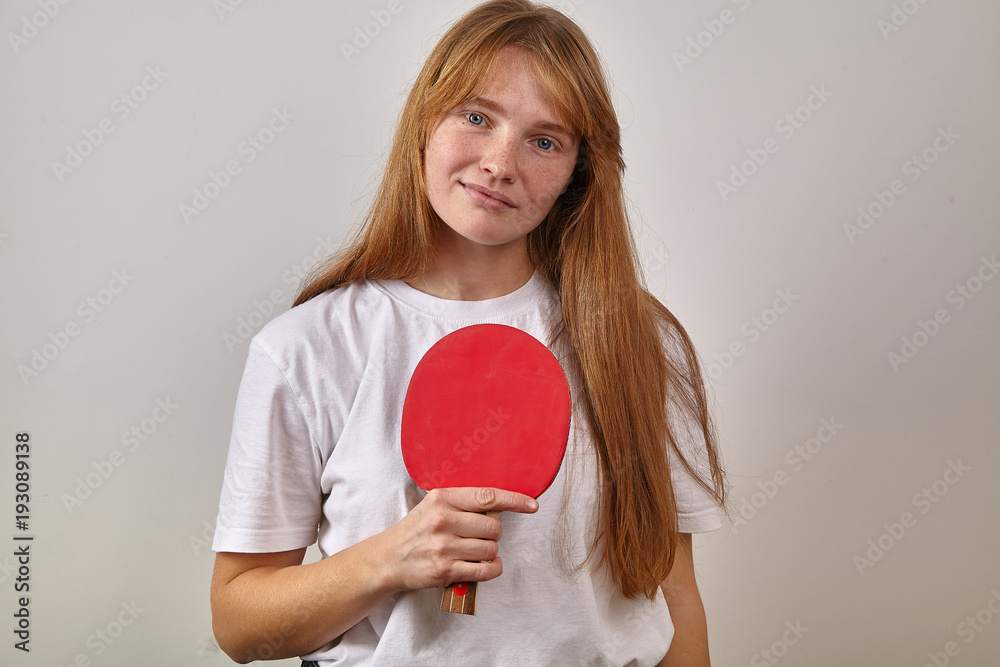Studio indoor portrait of young girl with red hair and freckles dressed in white t-shirt holding table tennis racket and looking at the camera Studio indoor portrait of young girl with red hair and freckles dressed in white t-shirt holding table tennis racket and looking at the camera