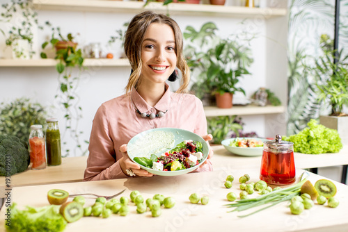Portrait of a young woman holding a plate of salad sitting indoors surrounded with green flowers and healthy vegan food