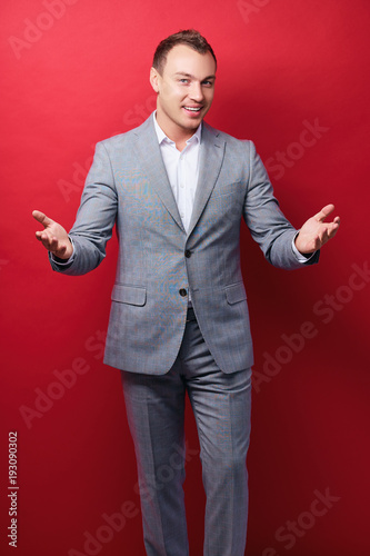 You are welcome! Cheerful young man in suit gesturing welcome sign and smiling while standing against red wall.