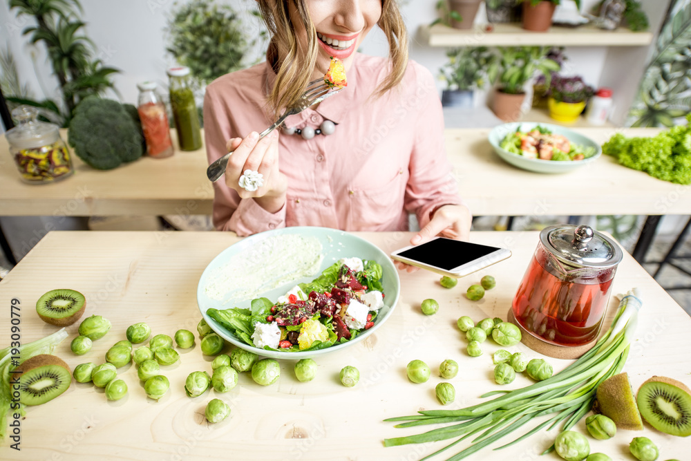 Plakat Woman eating salad and using smartphone at the table dwcorated with green food ingredients. Top view