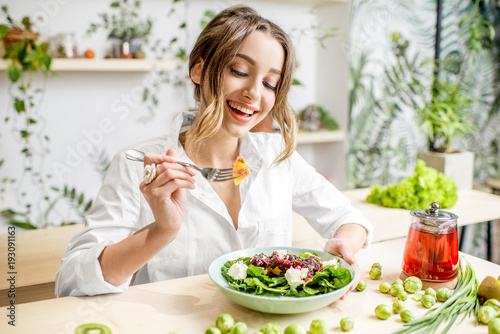Plakat Young woman eating healthy food sitting in the beautiful interior with green flowers on the background