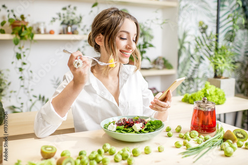 Young woman eating healthy food sitting with smartphone in the beautiful interior with green flowers on the background