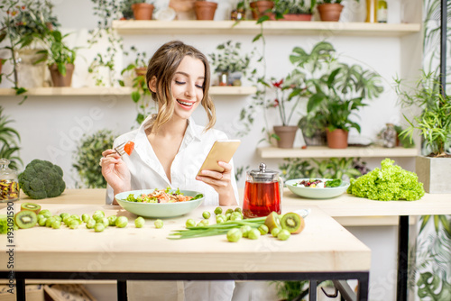 Young woman eating healthy food sitting with smartphone in the beautiful interior with green flowers on the background