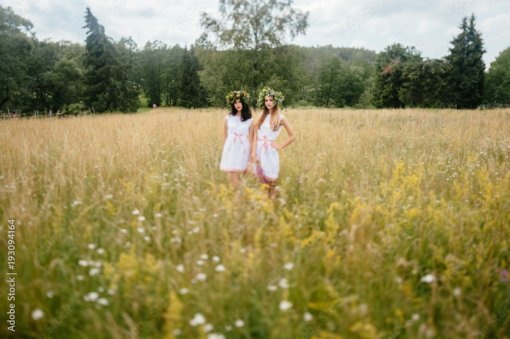 Loving couple at nature. Two pagan young girls of Slavonic appearance friendship. Women in traditional russian costumes. Feminine and tenderness. Lovely girls with wreath of flowers together in field. Loving couple at nature. Two pagan young girls of Slavonic appearance friendship. Women in traditional russian costumes. Feminine and tenderness. Lovely girls with wreath of flowers together in field.