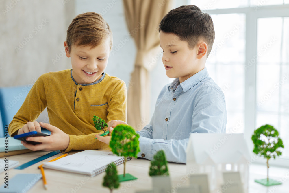 Curious kids. Pleasant cheerful pre-teen boys sitting at the table in the office and examining the workplace of their father, scrutinizing his models of landscape elements Curious kids. Pleasant cheerful pre-teen boys sitting at the table in the office and examining the workplace of their father, scrutinizing his models of landscape elements
