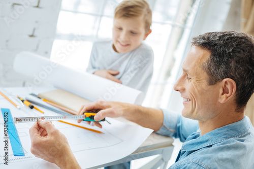 Important measures. Pleasant pre-ten boy sitting at the table in his fathers office and watching him check the measures on the tape while working on the blueprint