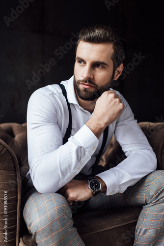 stylish man in suspenders thinking and sitting in armchair