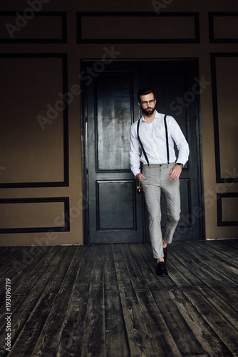 stylish elegant man posing in trendy suspenders against door in loft interior