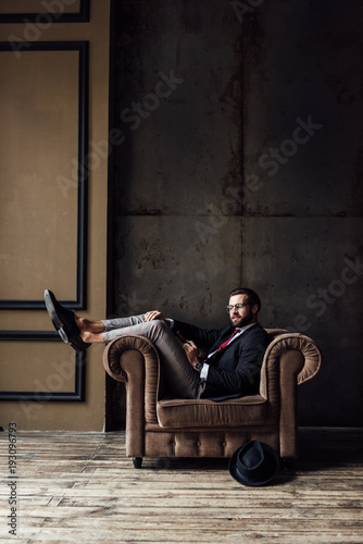 elegant businessman posing in armchair, hat lying on floor near, loft interior © LIGHTFIELD STUDIOS