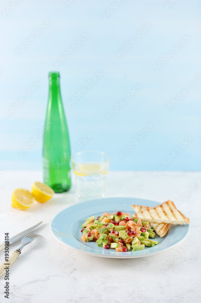 Tar-tar from salmon with avocado and pomegranat on a blue plate. On a plate toast. There are cutlery nearby. In the background, a lemon, a glass of water and a bottle. Light background. Vertical shot.