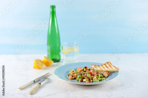 Tar-tar from salmon with avocado and pomegranate seeds on a blue plate. On a plate toast. There are cutlery nearby. In the background, a lemon, a glass of water and a bottle. Light background. 