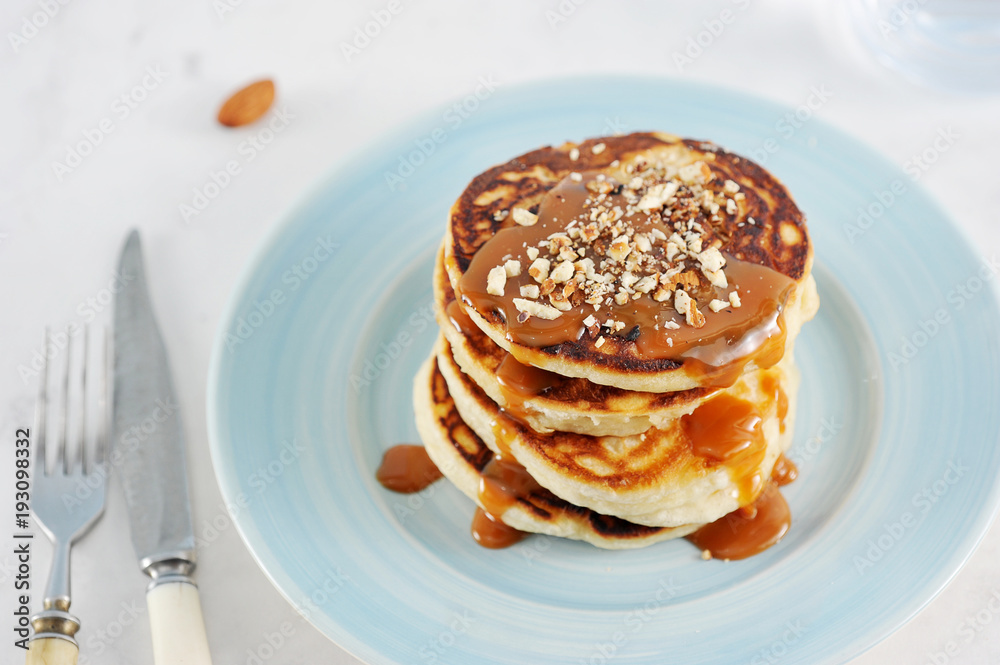 Pancakes with caramel and nuts. Several pancakes are lined with a pile, poured with caramel syrup, sprinkled with nutmeg almonds. Next to the plate are cutlery. Close-up. 