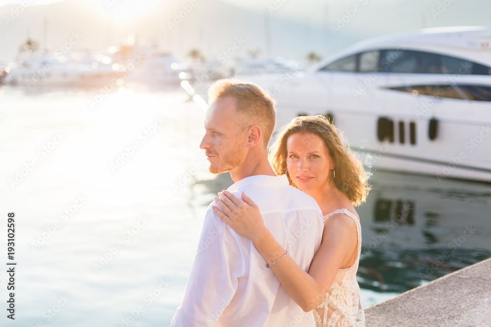 Couple of travelers are sitting in sea resort near yachts during romantic honeymoon vacation at sunset after wedding. Man and woman are happy tourists. Lovers are wearing in white clothes outdoor. Couple of travelers are sitting in sea resort near yachts during romantic honeymoon vacation at sunset after wedding. Man and woman are happy tourists. Lovers are wearing in white clothes outdoor.