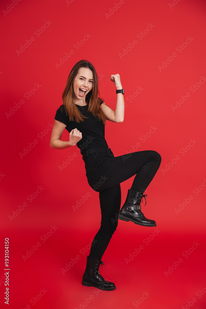 Full-length portrait of happy woman 20s wearing black t-shirt screaming and clenching fists in rejoicing victory or triumph, isolated over red background