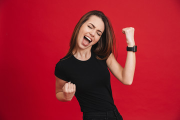 Portrait of ecstatic woman 20s wearing black t-shirt screaming and clenching fists like rejoicing victory or triumph, isolated over red background