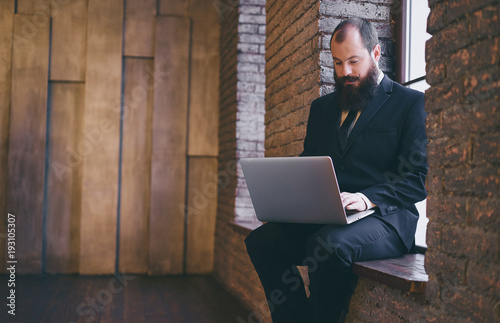 Workplace. Young bearded man wearing suit and tie using laptop while sitting near the window. photo