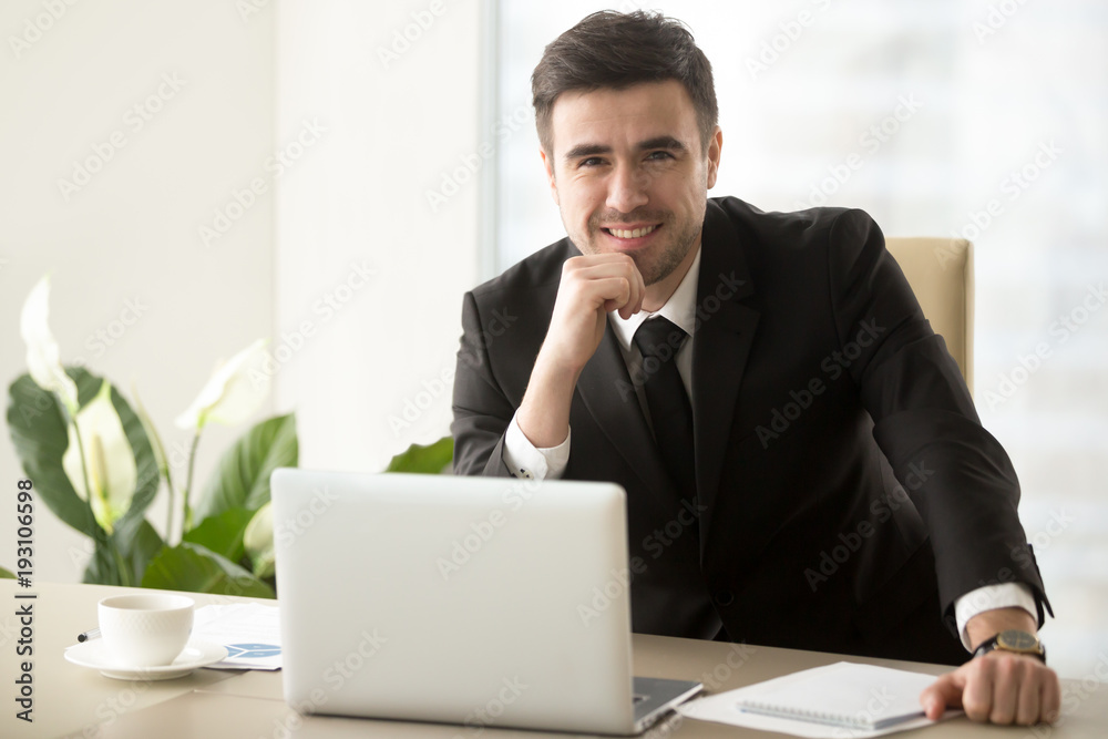Portrait of happy smiling businessman sitting at work desk in front of laptop, looking in camera. Successful attractive company executive manager posing at workplace, feeling confident and satisfied Portrait of happy smiling businessman sitting at work desk in front of laptop, looking in camera. Successful attractive company executive manager posing at workplace, feeling confident and satisfied