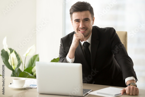 Portrait of happy smiling businessman sitting at work desk in front of laptop, looking in camera. Successful attractive company executive manager posing at workplace, feeling confident and satisfied