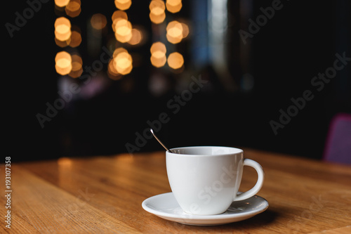 A white coffee cup with a spoon inside on a wooden table of a cafe  bokeh lights in the background.