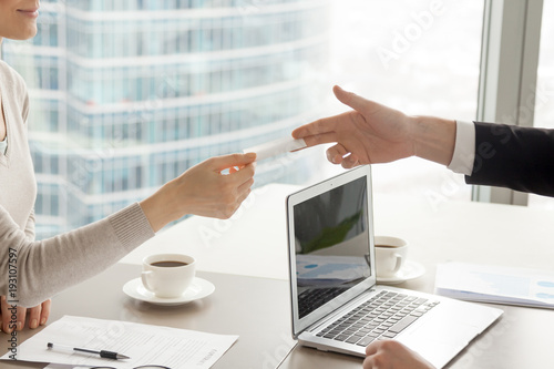 Businessman stretching out hand with blank business card over desk with laptop, coffee cups and documents to satisfied businesswoman after successful contract negotiation in company office. Close up