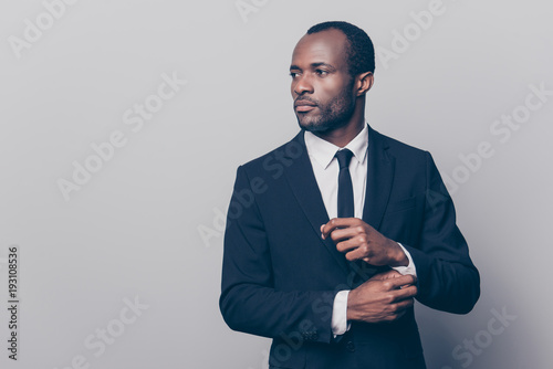 Portrait of stunning, trendy, attractive, dreamy, perfect man in black suit with tie fasten button on sleeve cuffs of white shirt, looking to the side, isolated on grey background