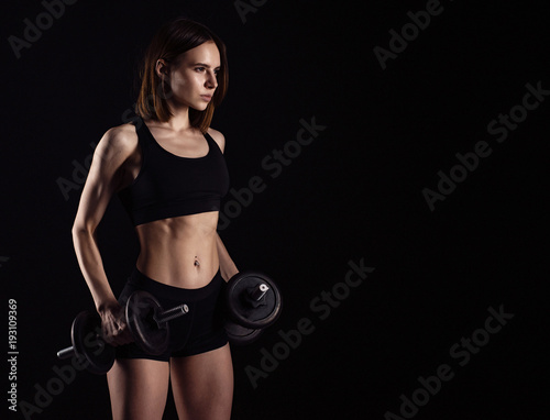Sporty muscular female doing workout with dumbbells isolated on black background. Athletic young woman do a fitness workout with weights.