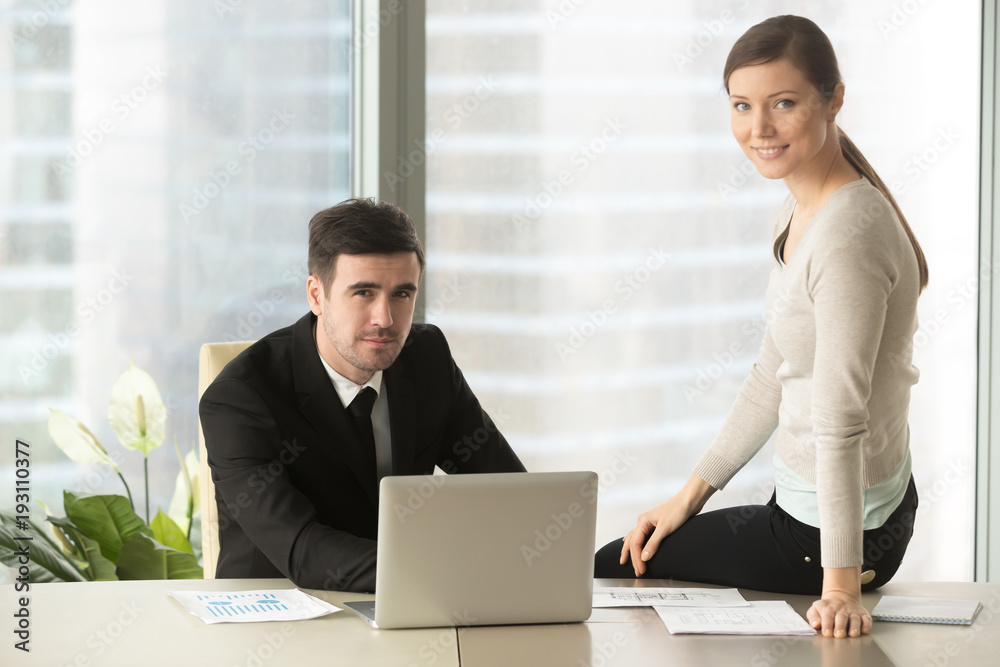 Candid portrait of millennial male and female business leaders sitting together at office desk with laptop and papers and looking at camera with smile. Positive company coworkers posing at workplace Candid portrait of millennial male and female business leaders sitting together at office desk with laptop and papers and looking at camera with smile. Positive company coworkers posing at workplace