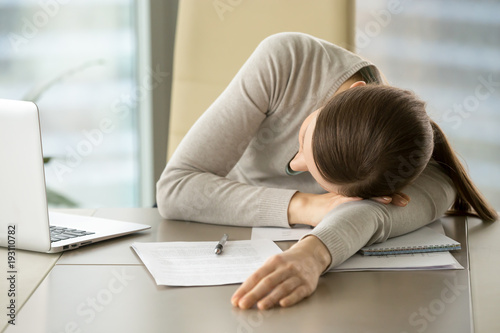 Tired female office worker lying on desk with laptop and business documents because of overwork. Exhausted from hard paperwork businesswoman sleeping on workplace, taking short napping during work day photo