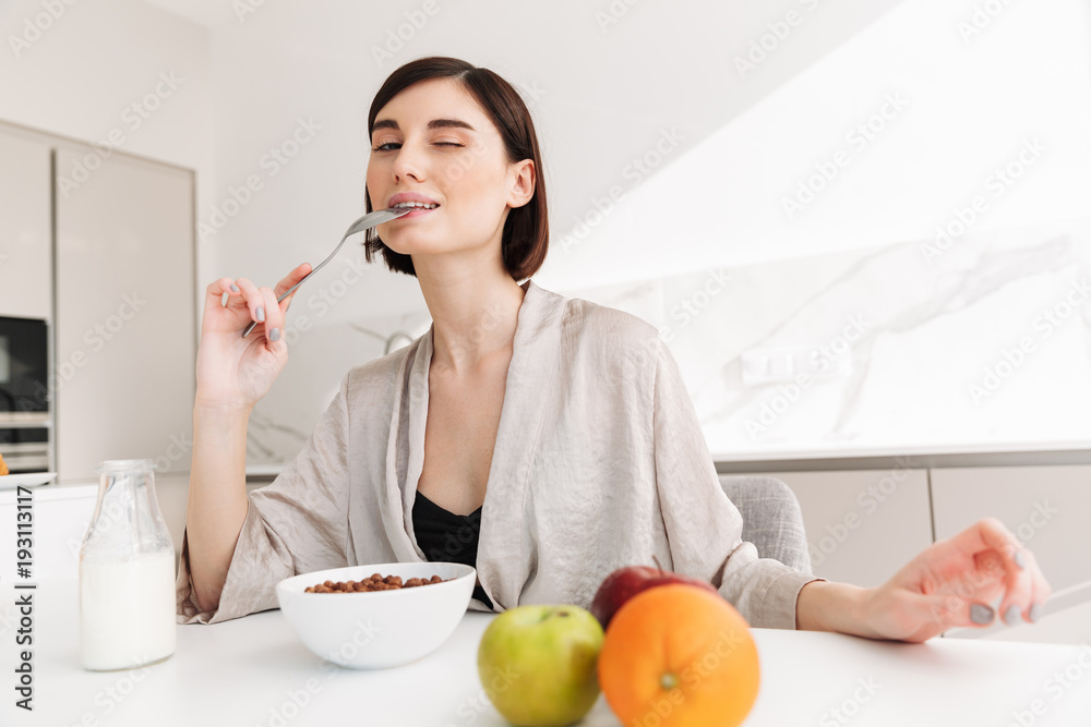 Photo of joyful young woman having breakfast in kitchen of hotel apartment, while eating cocoa balls with milk and fruits Photo of joyful young woman having breakfast in kitchen of hotel apartment, while eating cocoa balls with milk and fruits