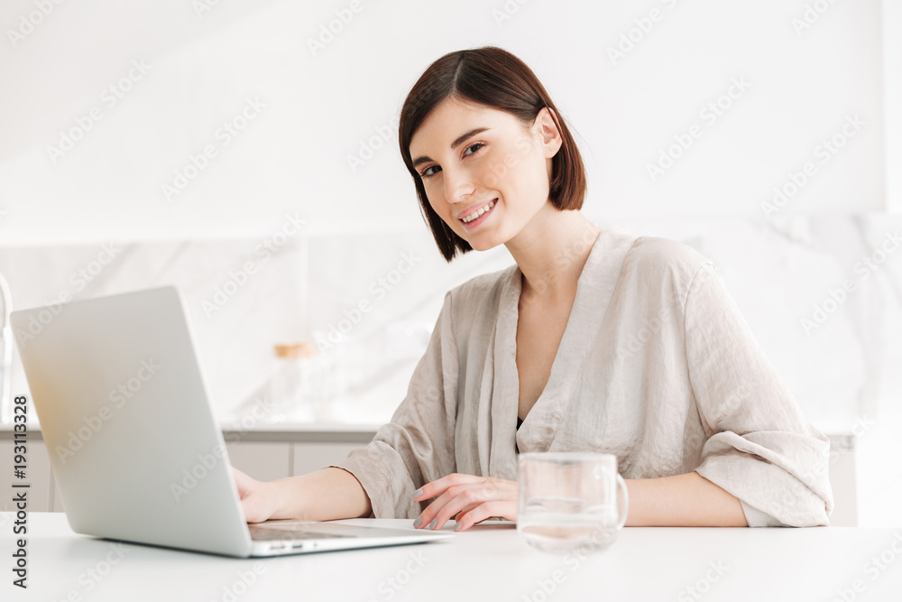 Photo of beautiful caucasian woman wearing robe looking on camera with smile while working, or communicating on white notebook at home Photo of beautiful caucasian woman wearing robe looking on camera with smile while working, or communicating on white notebook at home