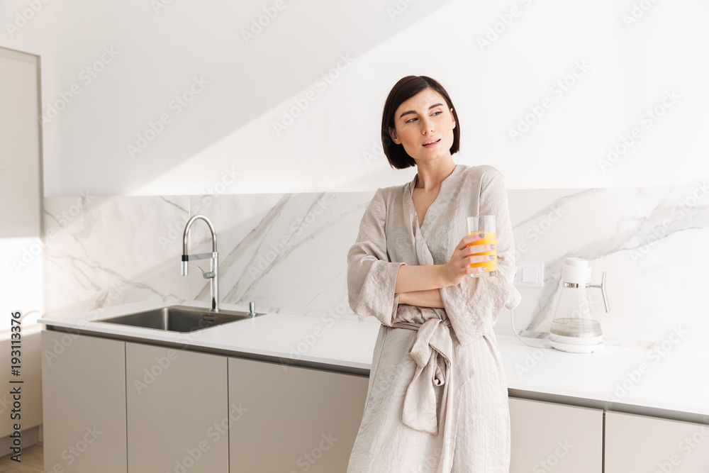 Young fascinating woman with short dark hair standing in kitchen and drinking fresh orange juice, from transparent glass Young fascinating woman with short dark hair standing in kitchen and drinking fresh orange juice, from transparent glass