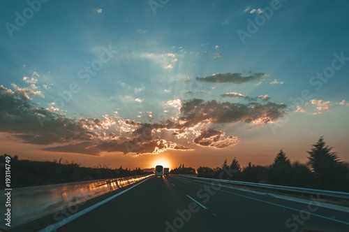 Colorful sunset over at highway. Landscape with orange sunset over highway. Cloud layers with sunrays between them. Contre-jour silhouette of cars driving on highway.