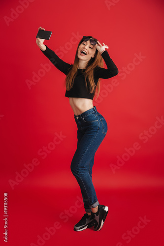 Full-length photo of splendid woman with long brown hair taking selfie on smartphone while touching sunglasses, isolated over red background