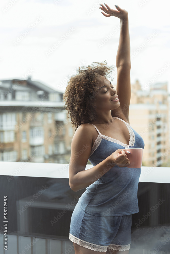 Cheerful young african woman tasting mug of appetizing tea while stretching on balcony