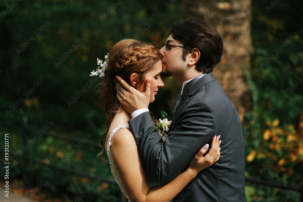 Groom kisses bride's forehead tender standing somewhere in the park Groom kisses bride's forehead tender standing somewhere in the park