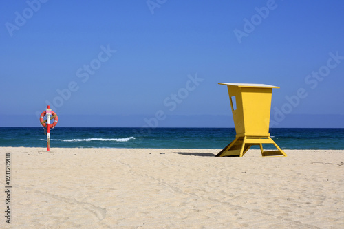 Gelber überdachter Sitz für Rettungsschwimmer und Rettungsreifen an hellem Sandstrand.