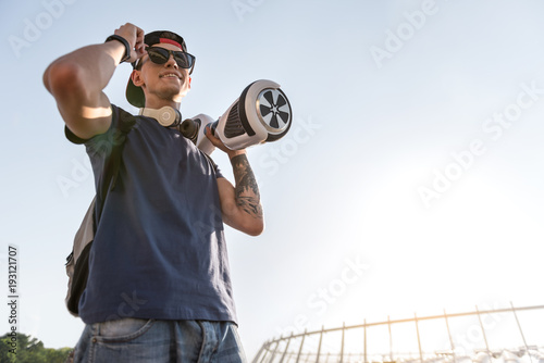 Low angle portrait of beaming teenager holding hoverboard at street. Technology concept. Copy space