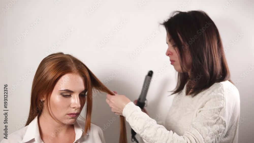 Stylist is making hairstyle for red haired woman. Master is holding curling tongs, taking strand of hair and curling it Stylist is making hairstyle for red haired woman. Master is holding curling tongs, taking strand of hair and curling it