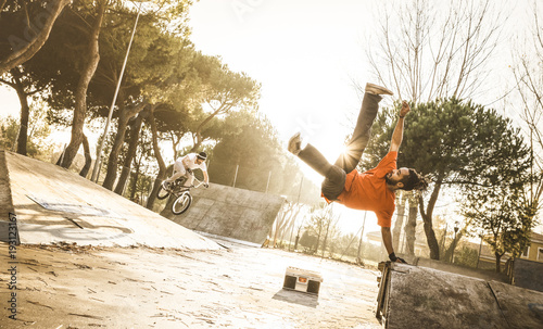 Urban athlete breakdancer performing acrobatic jump flip at skate park - Guy riding bmx bicycle behind mate acrobat dancing with extreme move - Breakdance and friendship concept - Warm sunset filter photo